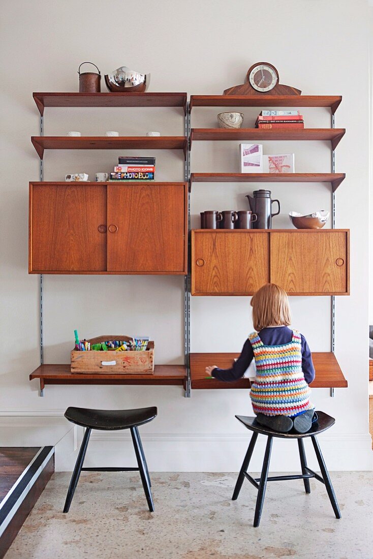 Little girl sitting on wooden stool in front of retro-style, modular shelving system with compartments, cabinets and writing desk on wall-mounted rails