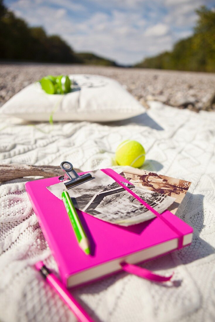 Notizbuch mit pinkfarbenem Einband und knallig gelber Tennisball auf naturfarbener Strickdecke im Freien