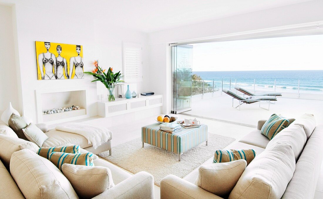Grosszügige Lounge mit naturfarbener Sofagarnitur und gepolsterter Couchtisch vor offener Terrassen-Falttür mit Panoramablick auf das Meer