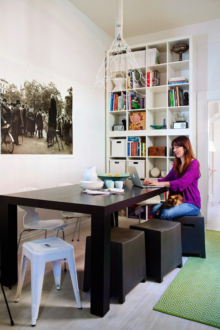 Frau mit Laptop an dunklem Holztisch, verschiedene Hocker im Stilmix, im Hintergrund Bücherregal