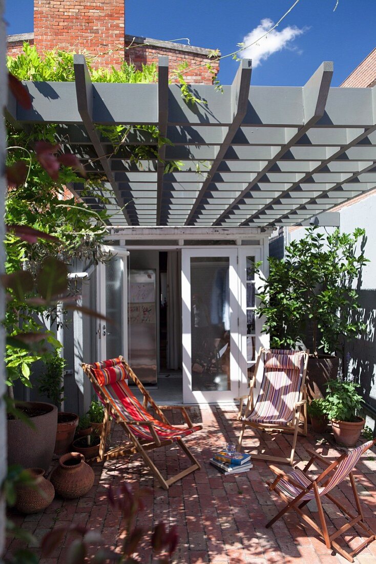 Deckchairs on sunny terrace below grey wooden lattice pergola adjoining house with open door