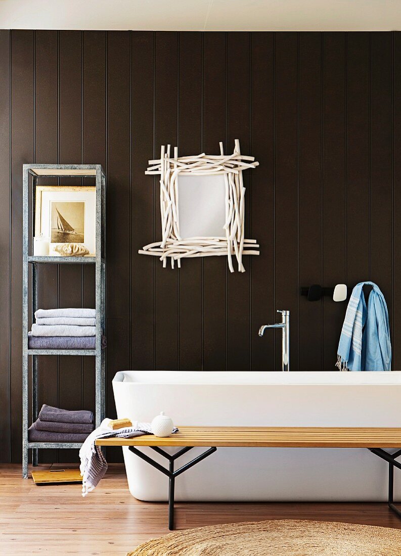 Bathroom display - simple wooden bench, bathtub, metal shelves and mirror framed with white sticks on dark brown wooden wall