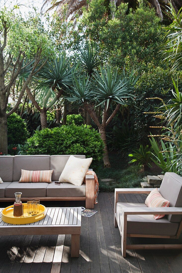 Grey-upholstered outdoor furniture and coffee table on wooden terrace with yuccas in mature garden in background