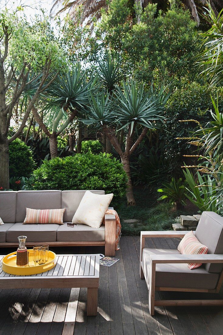 Holzterrasse mit hellgrauen Outdoor Polstermöbeln und Coffee-Table; im Hintergrund eingewachsener Garten mit Yucca Palmen