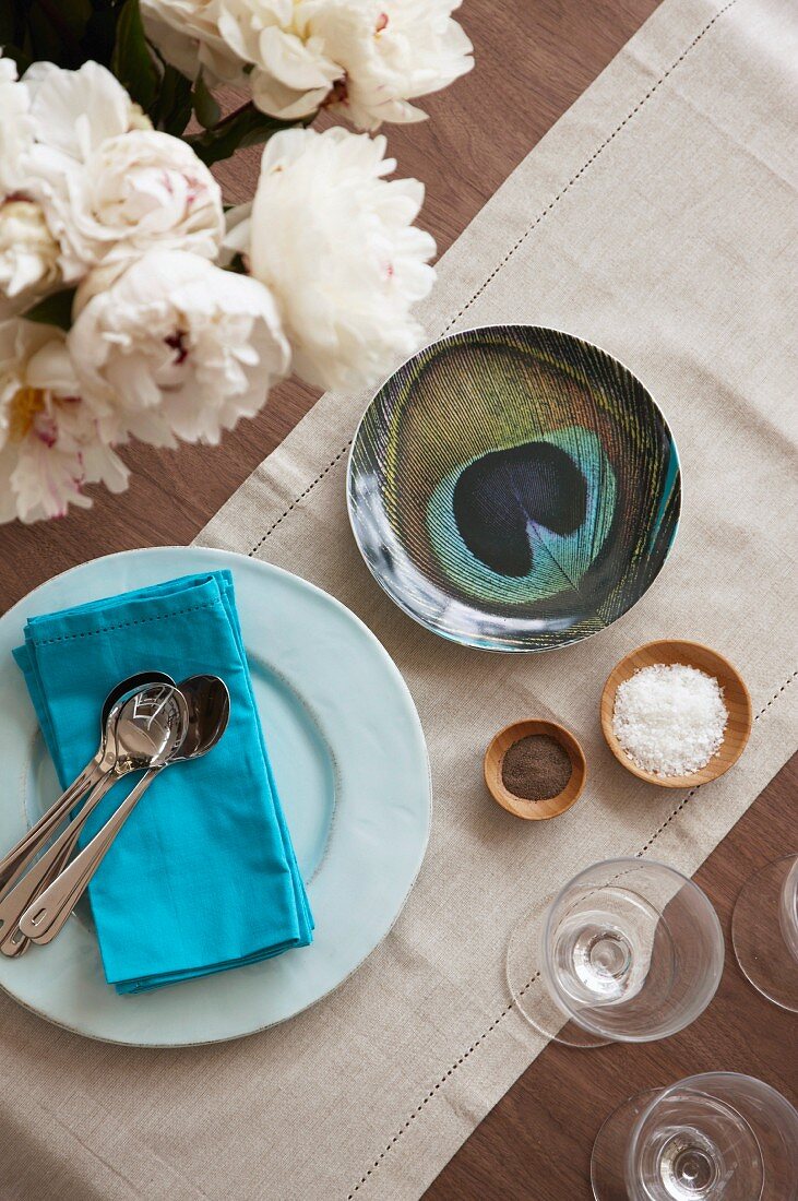 Teller mit Besteck auf blauer Serviette neben Schale mit Pfauenaugen Muster und Holzschälchen auf hellem Leinen Tischläufer