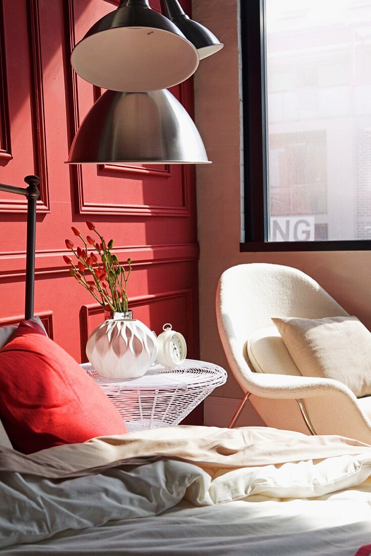 Rotes Kissen auf Bett, neben Drahtgeflecht-Beistelltisch und weisser Sessel am Fenster in Zimmerecke vor lachsrot lackierter Holz Kassettenwand