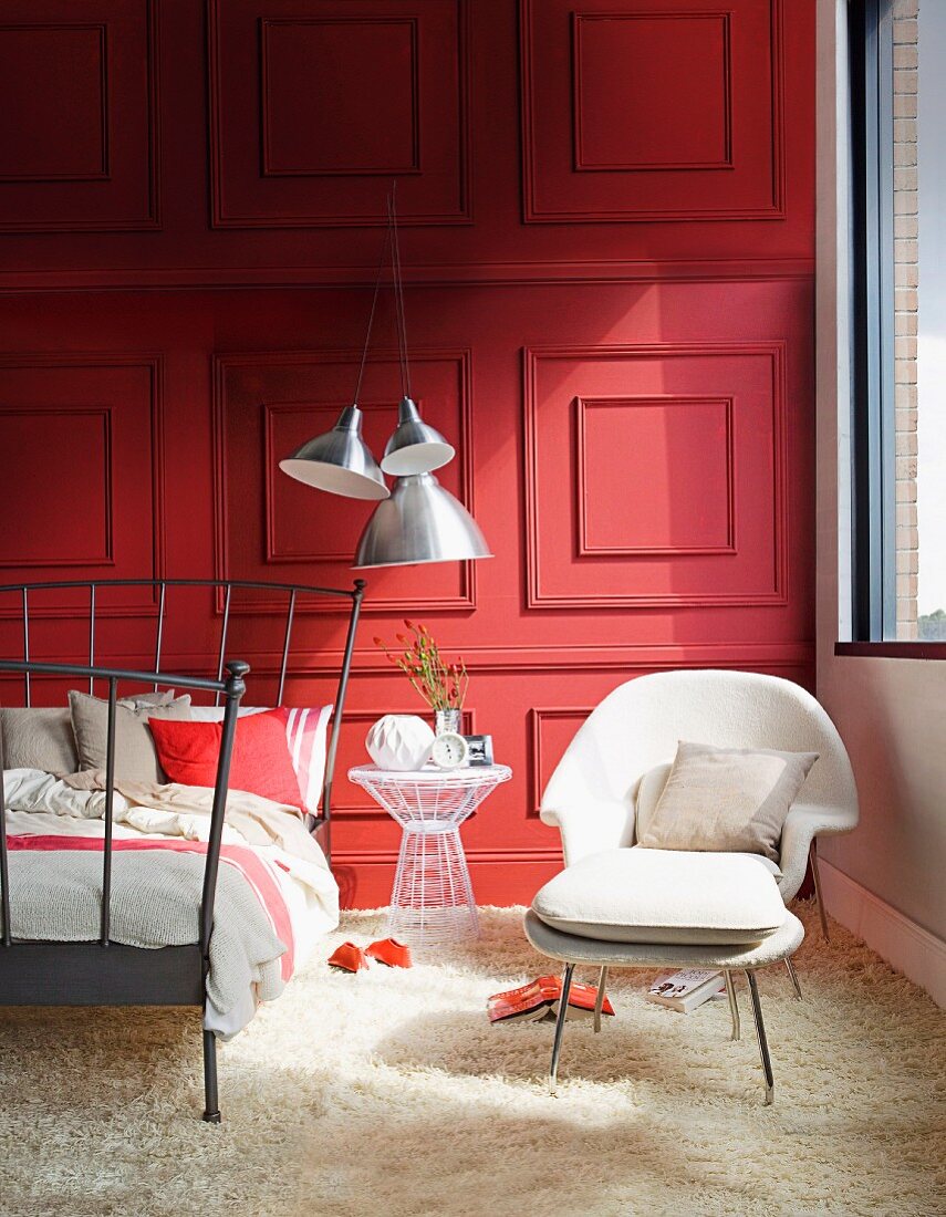 Dark grey metal bed frame, white armchair with matching footstool on white flokati rug in front of red-painted wood-panelled wall