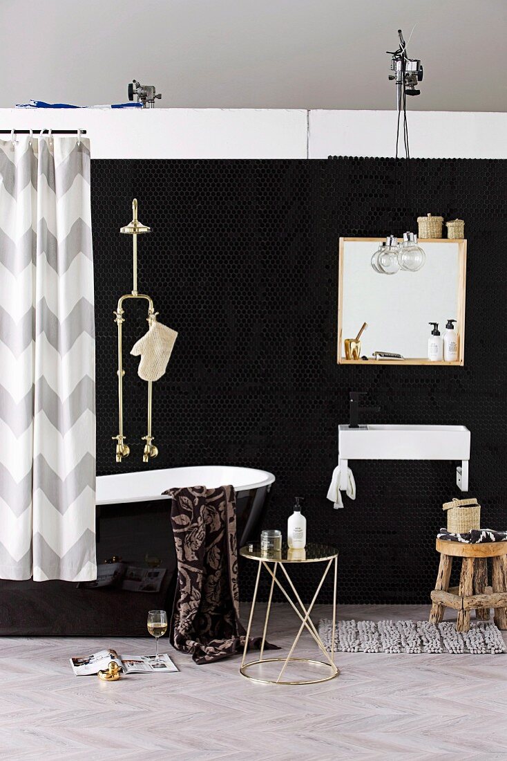 Badezimmer in Schwarz - Metall Beistelltisch vor schwarzer, glänzender Badewanne mit Duscharmatur und Spiegel über Waschbecken an schwarzer Wand