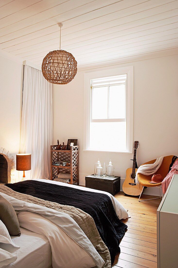 Double bed and wicker pendant lamp suspended from white wood-clad ceiling in simple bedroom with trunk and guitar next to retro armchair against wall