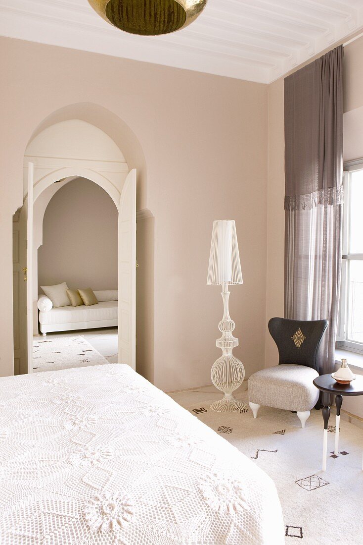 Schlafzimmer mit teilweise sichtbarem Bett, Stehlampe und Sessel am Fenster neben offener Tür mit Blick in Loungebereich