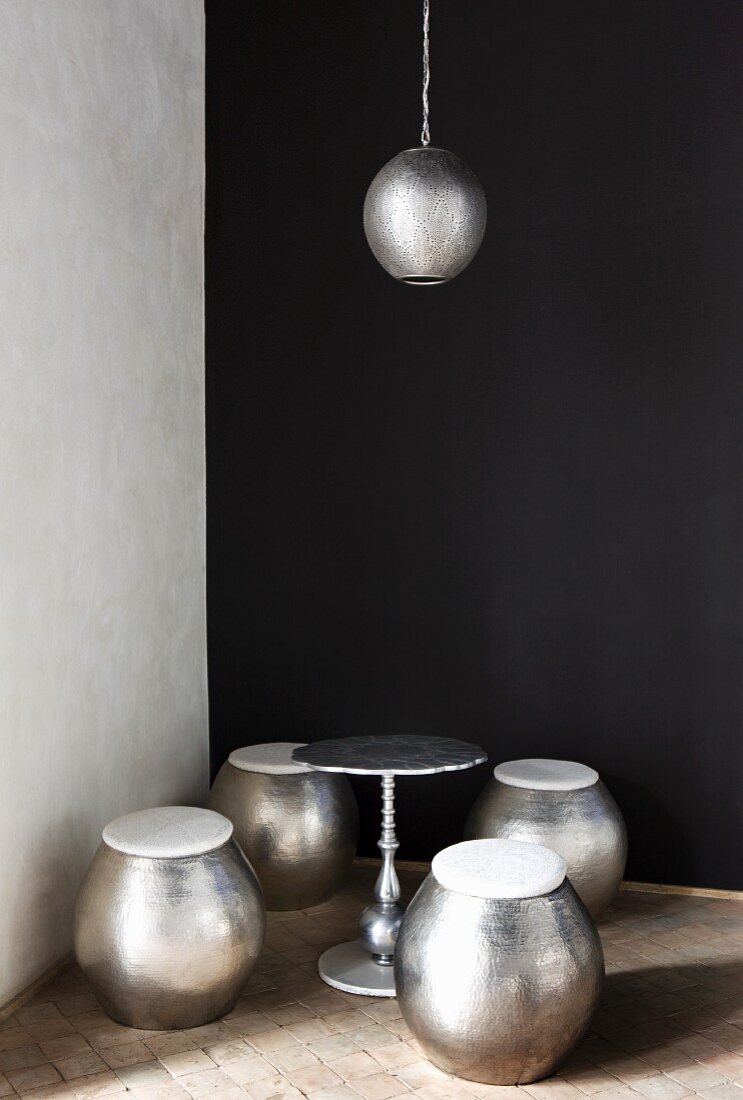 Bauchige Sitzhocker aus Metall mit Beistelltisch & Hängeleuchte in Zimmerecke vor schwarzer Wand