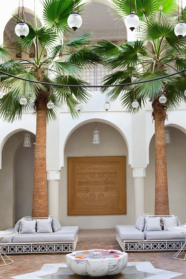 Marokkanischer Innenhof mit kreisförmiger Beleuchtung über Brunnen und Sitzgruppen um Palmen vor Arkaden