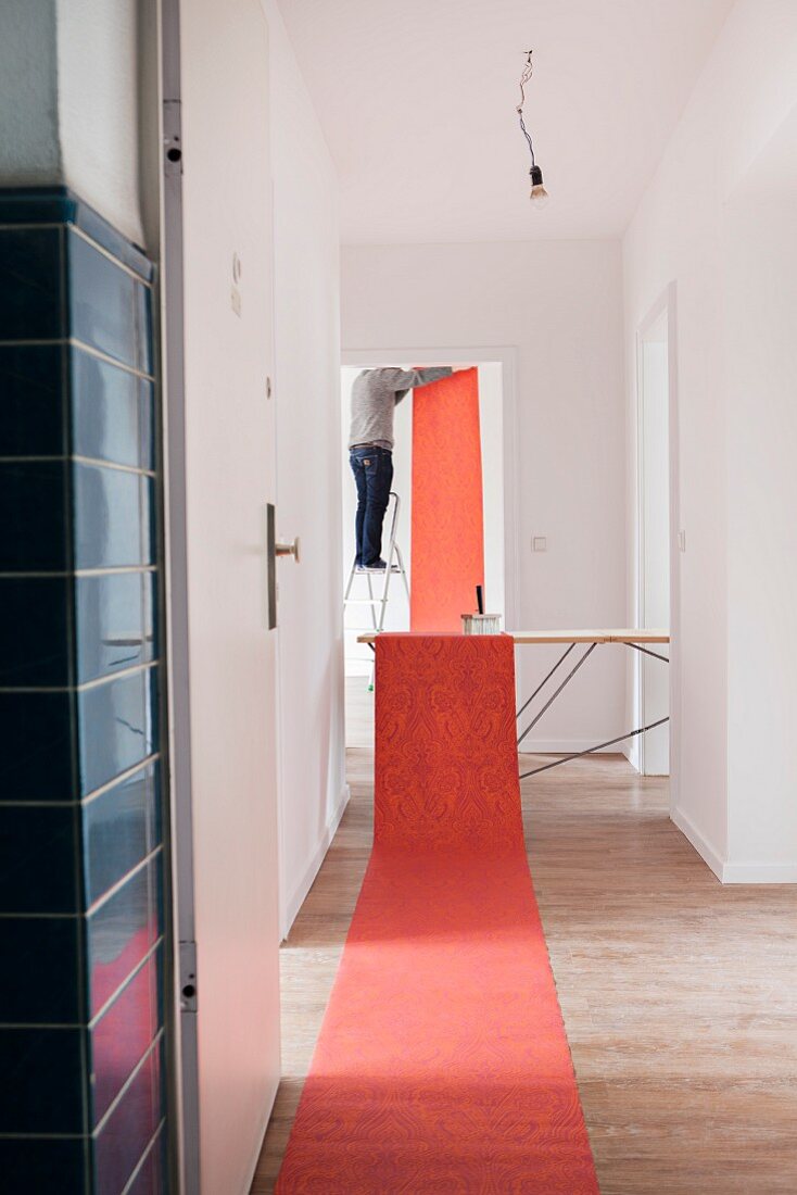 Rotgemusterte Tapete auf Boden und Tisch ausgerollt, im Hintergrund Blick auf Mann bei Tapezierarbeiten