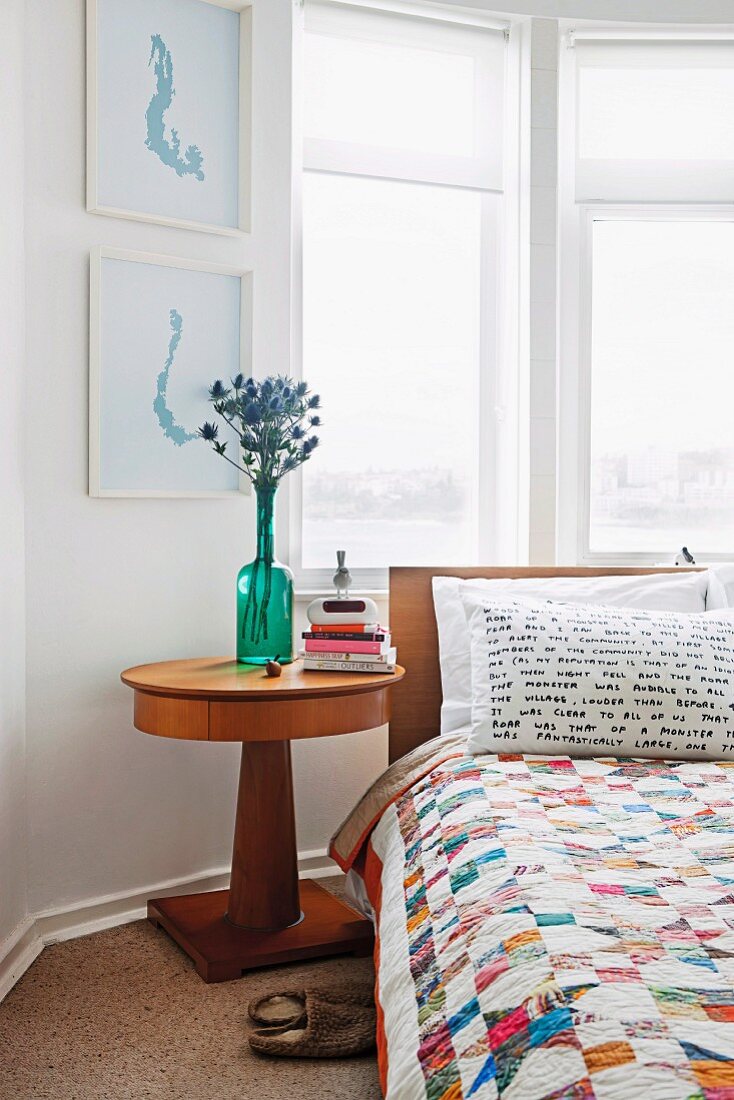 Schlafzimmer mit Bett, Patchworkdecke & Nachttisch mit runder Holzplatte vor Erkerfenster