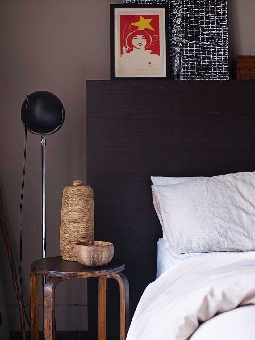 Teilweise sichtbares Bett mit dunklem Kopfteil neben schlichtem Hocker mit rustikalen Gefässen und Stehleuchte vor grauer Wand