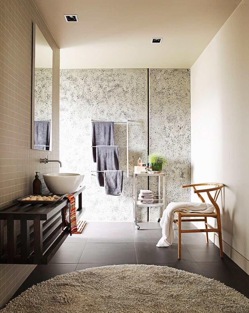 Modernes Badezimmer mit rundem Teppich auf schwarzem Fliesenboden, an Wand montierter Waschtisch & Klassiker-Stuhl aus Holz