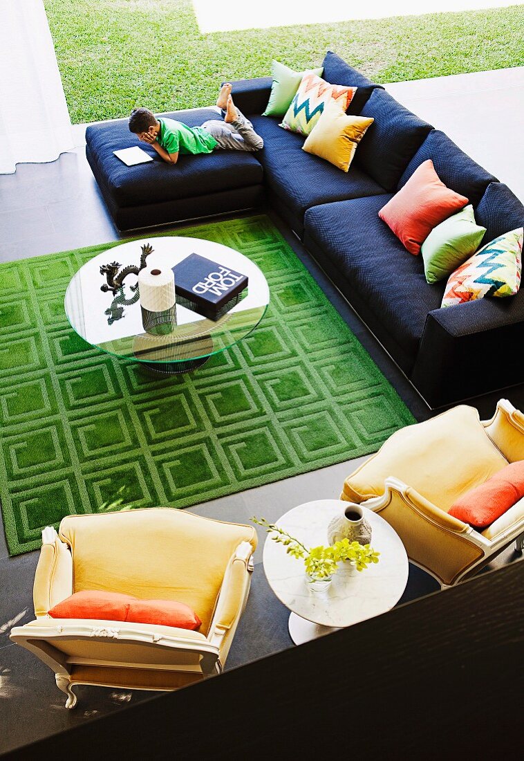 Blick auf grosszügigen Sitzbereich mit rundem Glastisch, grünem Teppich, moderner Couch & gelben Sesseln im Neo-Rokokostil