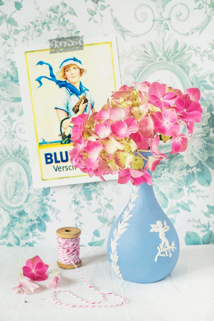 Pink hydrangea in vase next to spool of yarn on surface in front of vintage poster on blue and white wallpaper
