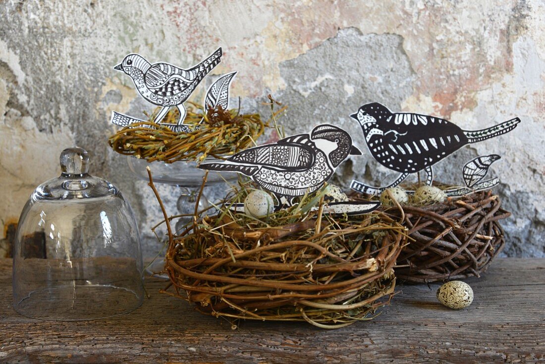 Hand-crafted paper birds and quail eggs in birds' nests on wooden surface against wall with peeling paint