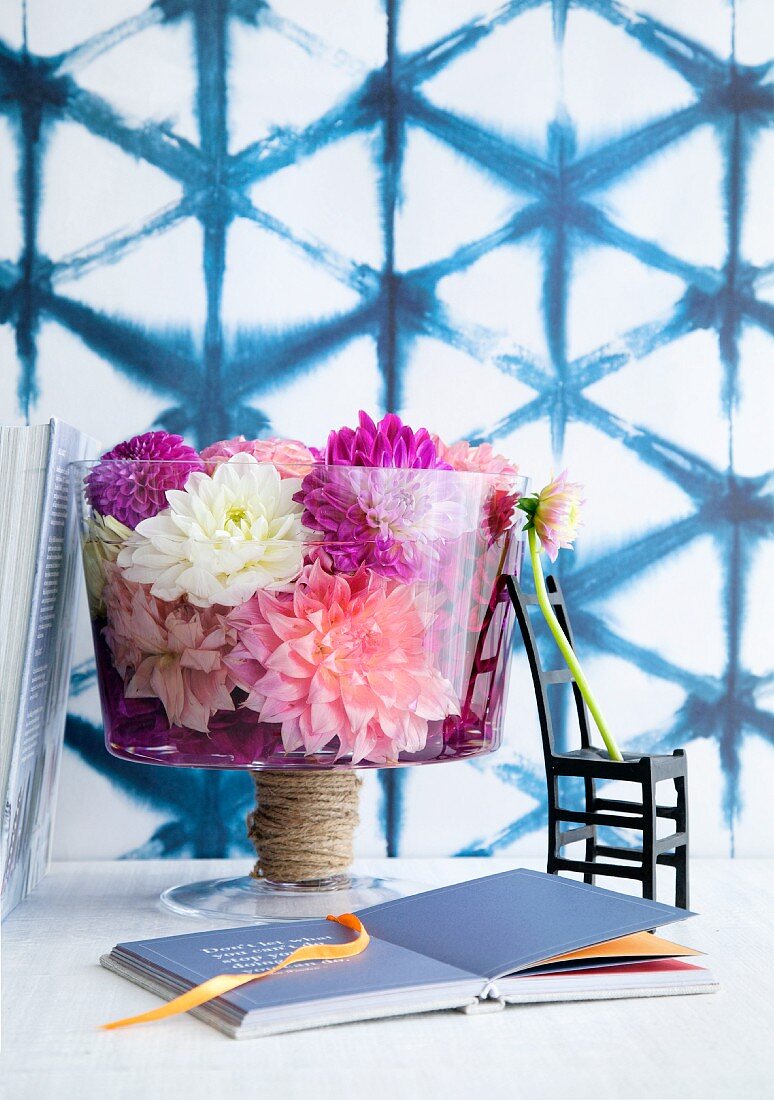 Glasschale mit Dahlienblüten, schwarzes Dekostühlchen und Buch vor Wand mit blauer Gitterstruktur