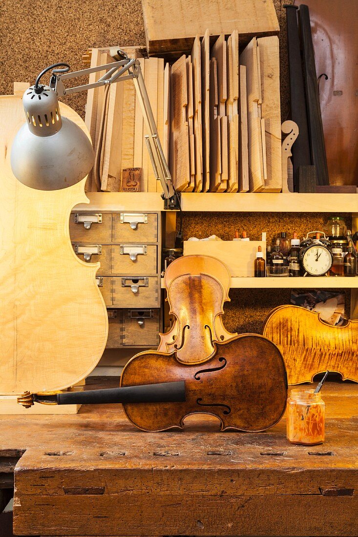 Unstrung violin amongst further string instruments on rustic workbench and assorted wood on shelf lit by retro desk lamp
