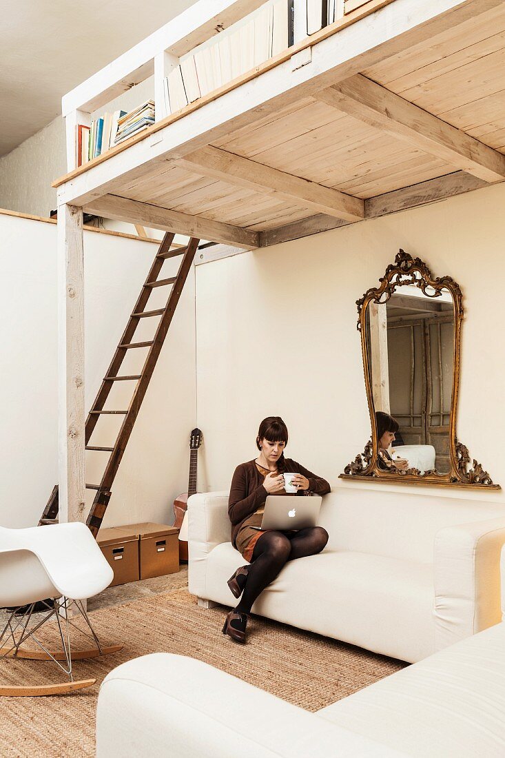 Junge Frau mit Laptop auf ecru-farbener Polstercouch unter weiß gestrichener Holzgalerie mit angelehnter Holzleiter in offenem Wohnraum mit Vintage-Flair