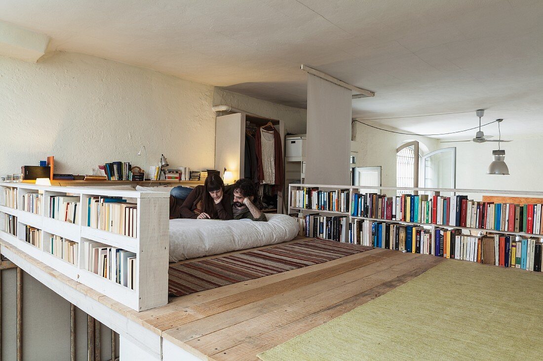 Junges Paar auf Holzgalerie lesend, umrahmt von Büchern in Bücherregalen als Galeriebrüstung