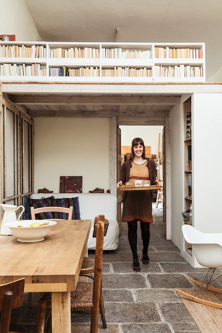 Junge Frau mit Tablett unter weiß gestrichener Holzgalerie in offenem Wohnraum mit Vintage-Flair und Stauraumlösung für Bücher