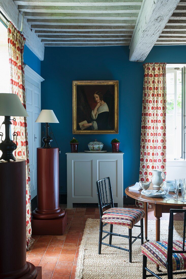 Round table and white, half-height cabinet below portrait of woman on blue wall in rustic dining room with wood-beamed ceiling
