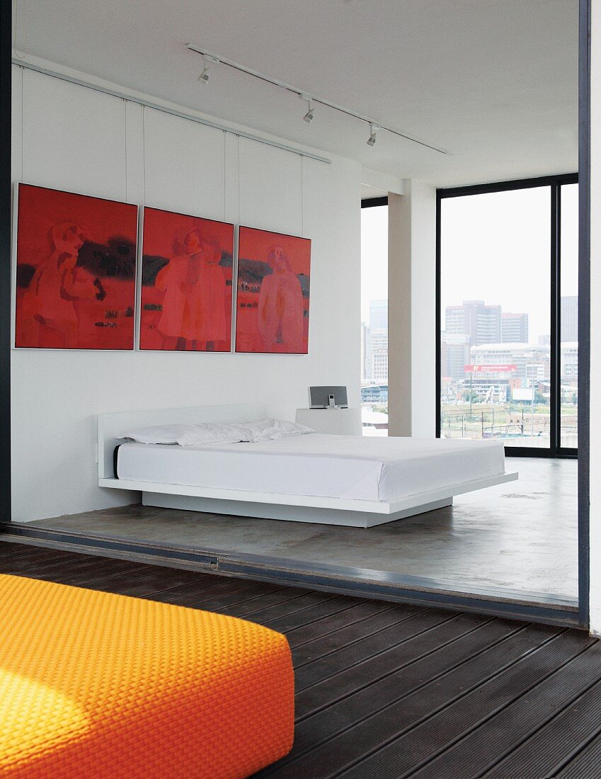 Red triptych above pedestal bed in loft apartment; view of Johannesburg cityscape through glass wall