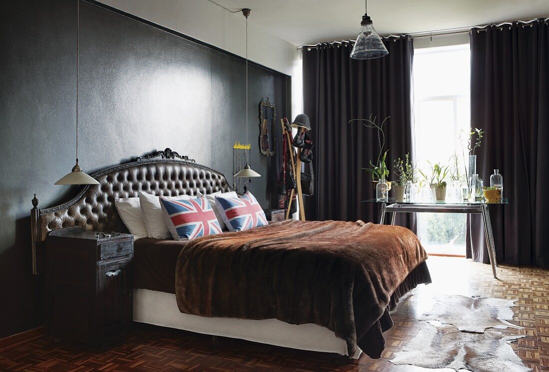 Doppelbett mit antikem Kopfteil, darauf Kissen mit Union-Jack-Motiv und Fell Tagesdecke, im Schlafzimmer mit dunkel getönter Wand und dunkelgrauen Vorhängen