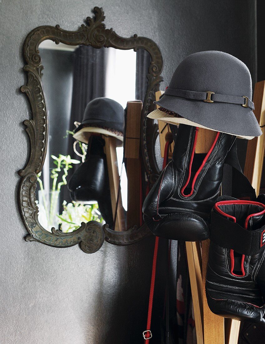 Hat and boxing gloves hanging on coat stand in front of mirror with ornate metal frame on grey-painted wall
