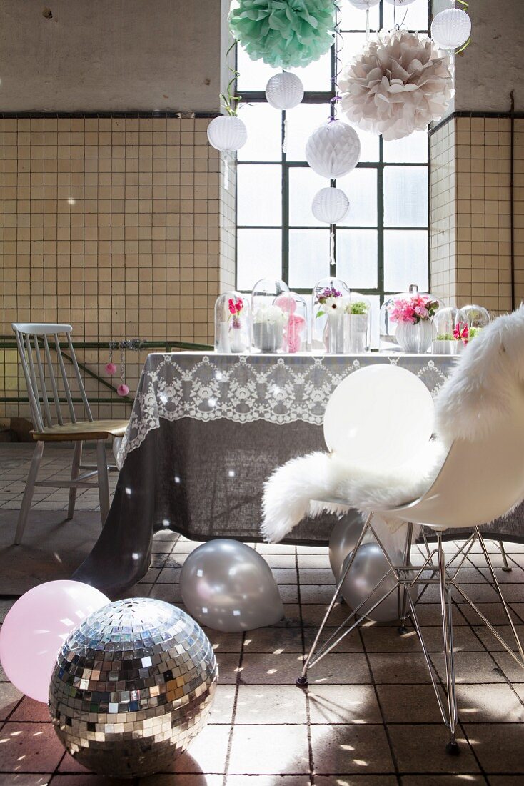 Feminine, romantische Partydekoration mit Lichteffekten von Discokugel, hängenden Pompons und Blumendeko auf Esstisch in Fabrikhalle mit Industriefenstern