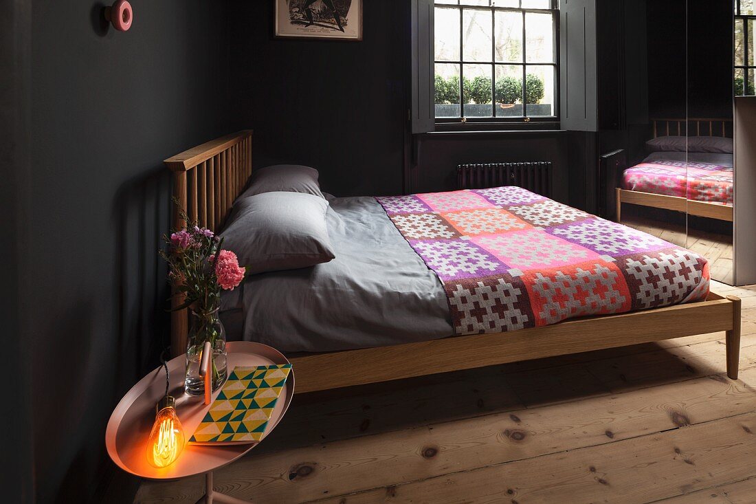 Bedroom with black-painted walls, wooden bed, colourful bedspread, mirrored wardrobe; lit lamp and vase of roses on tray table