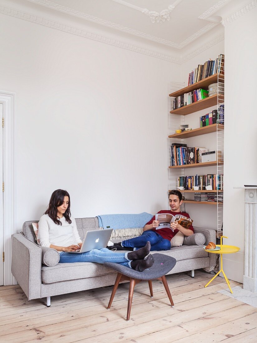 Young couple relaxing with magazine and laptop on grey sofa in renovated period apartment with wooden floor, bookcase and stucco ceiling