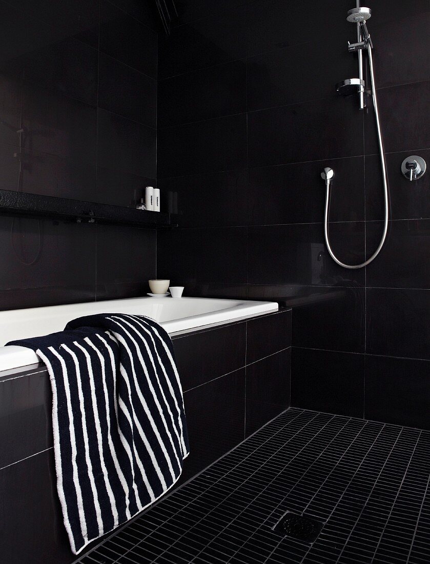 Black and white striped towel on edge of bathtub in black-tiled designer bathroom