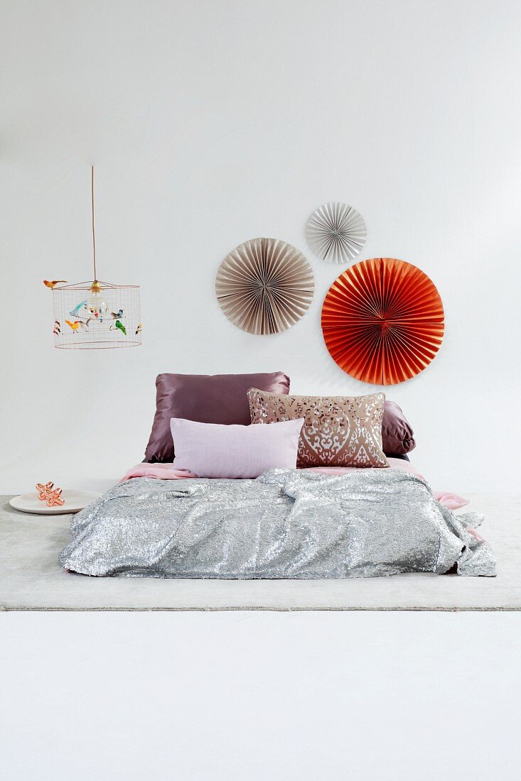 Shimmering combination of textiles on futon in front of decorative paper fans on wall
