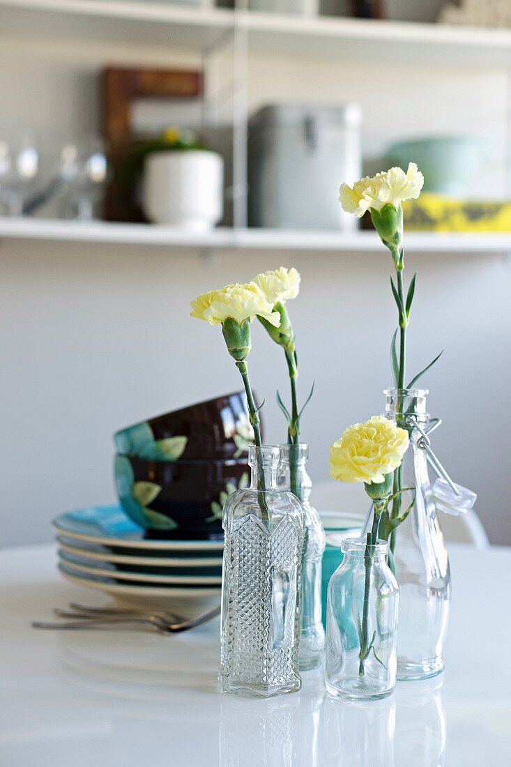 Gelbe Nelken in verschiedenen Glasgefässen vor einem Stapel farbigen Geschirrs auf einem Tisch