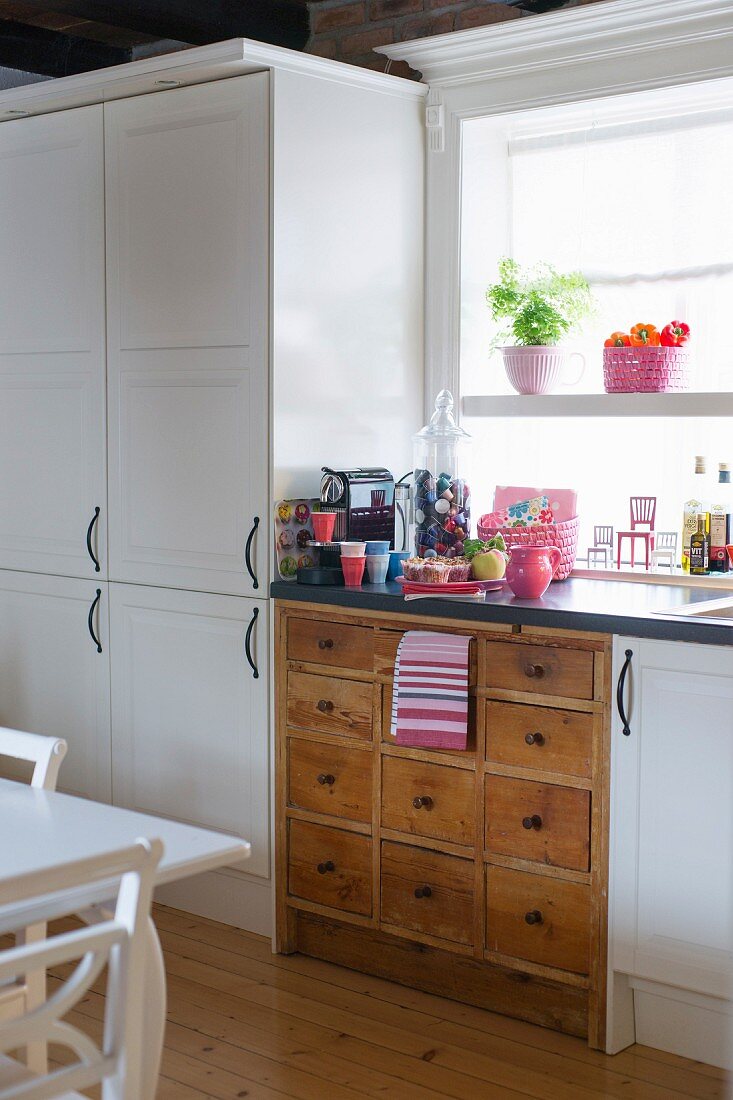 In eine weiße, moderne Küchenzeile integrierter, alter Schubladenkasten und rosarote Deko im Fenster