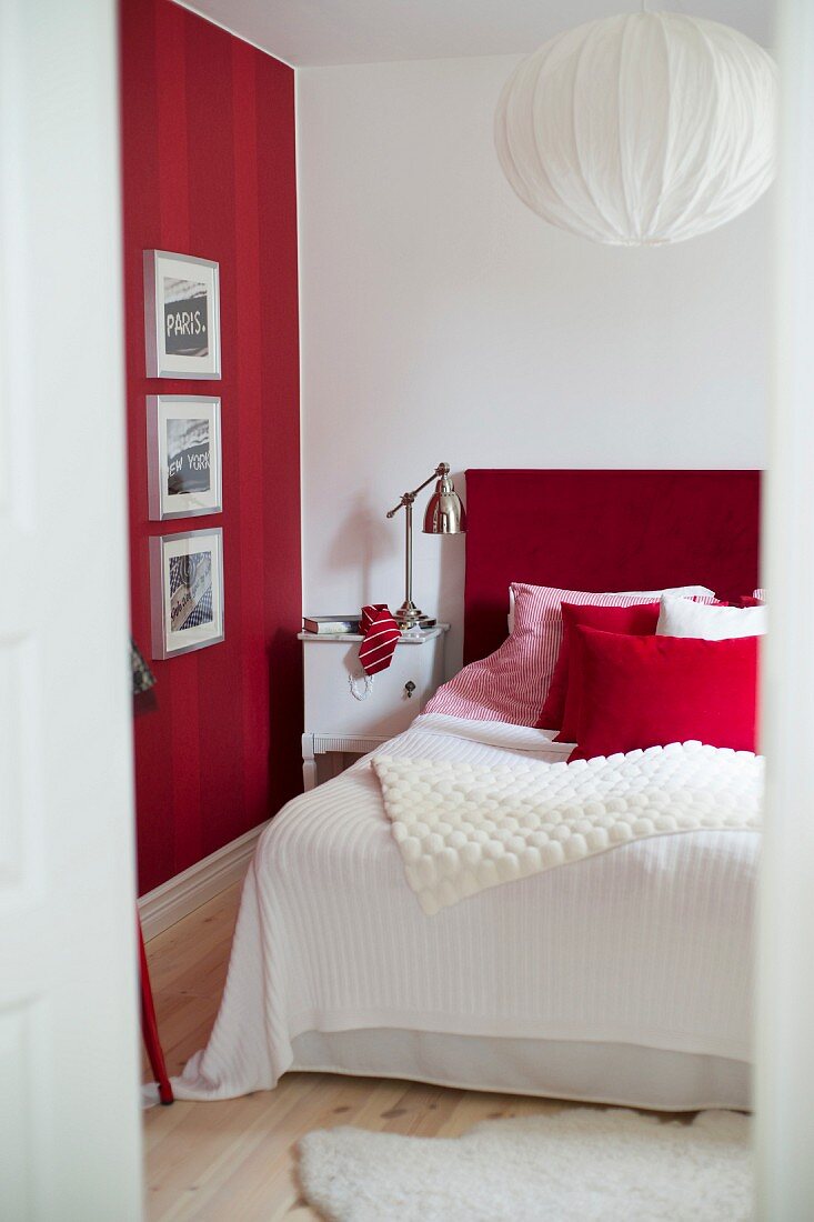 Gerahmte Bilder an rot gestreifter Wand, daneben ein Bett mit rotem Kopfteil und Leuchte im Retrostil auf Nachtkasten