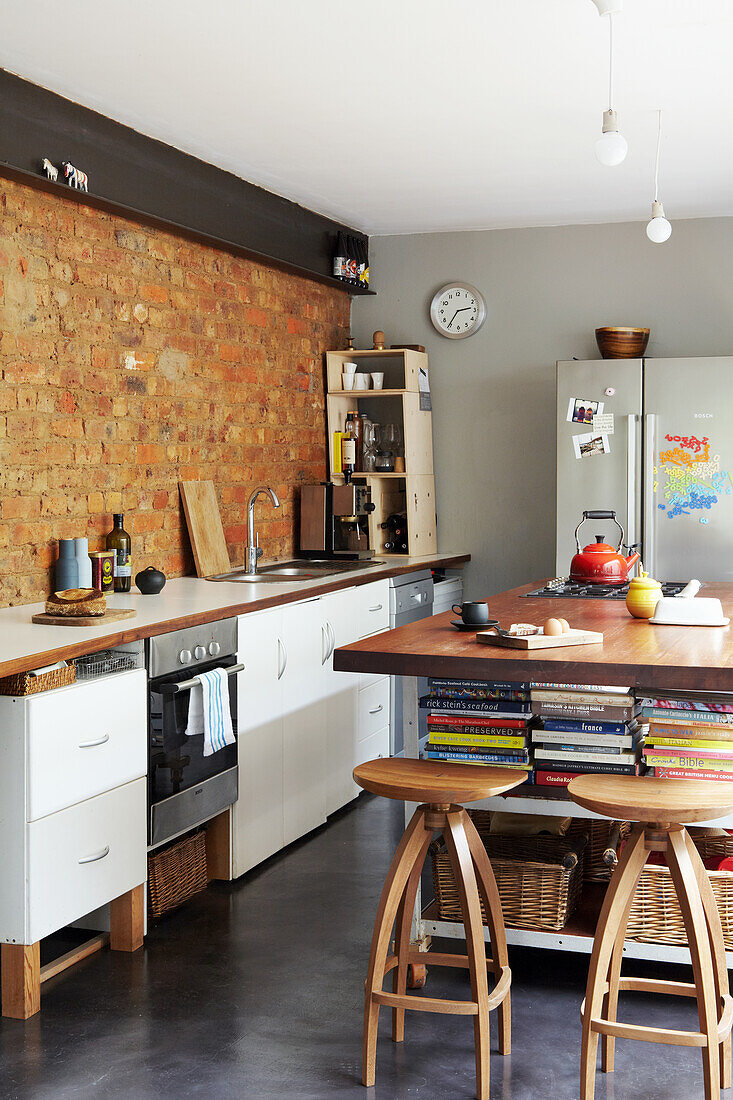 Kitchen unit in front of brick wall and kitchen island with storage space for books and vintage bar stools