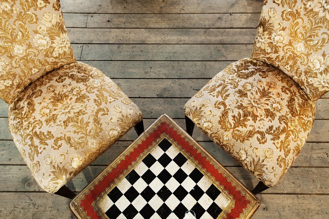 Table with integrated chess board and antique chairs with gold-patterned upholstery on rustic wooden floorboards