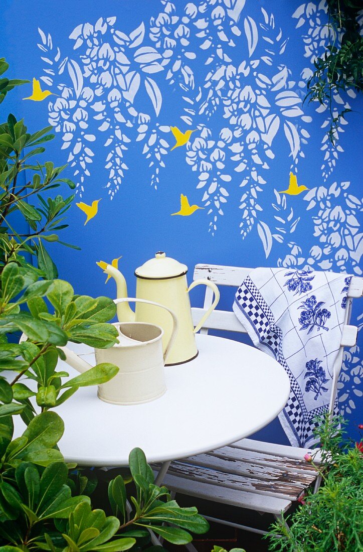 Wandbild mit Kolibris und Wysteria auf einer blau bemalten Gartenmauer, davor ein weißer Tisch und Stuhl mit Kaffeekanne