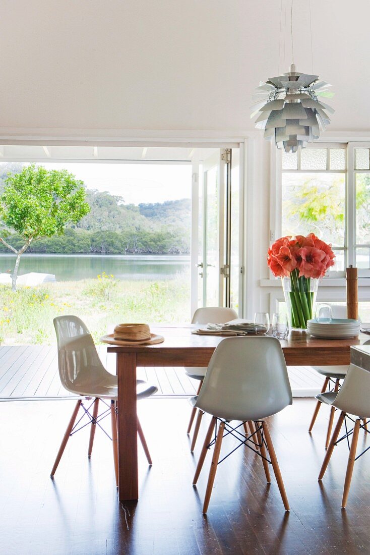 Classic, white shell chairs around wooden table in modern interior in front of open terrace door with view of lake