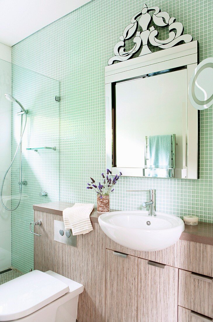 Waschtisch mit teilweise eingebautem Becken unter verziertem Spiegel an Mosaikfliesenwand, in der Ecke verglaster Duschbereich