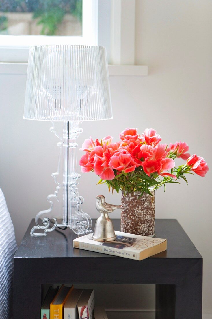 Roter Blumenstrauss in Vase neben Tischleuchte aus transparentem Kunststoff auf dunklem Nachttisch