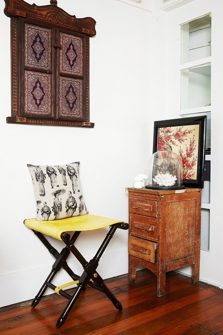 Folding stool in corner next to small, vintage cabinet & wooden objet d'art on wall