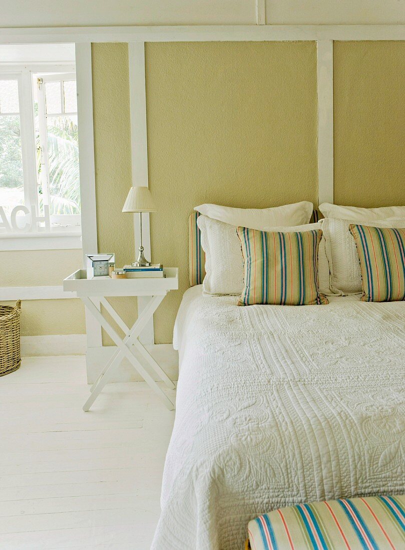 Double bed with scatter cushions and white bedspread in front of pale green wood-panelled wall in rustic atmosphere