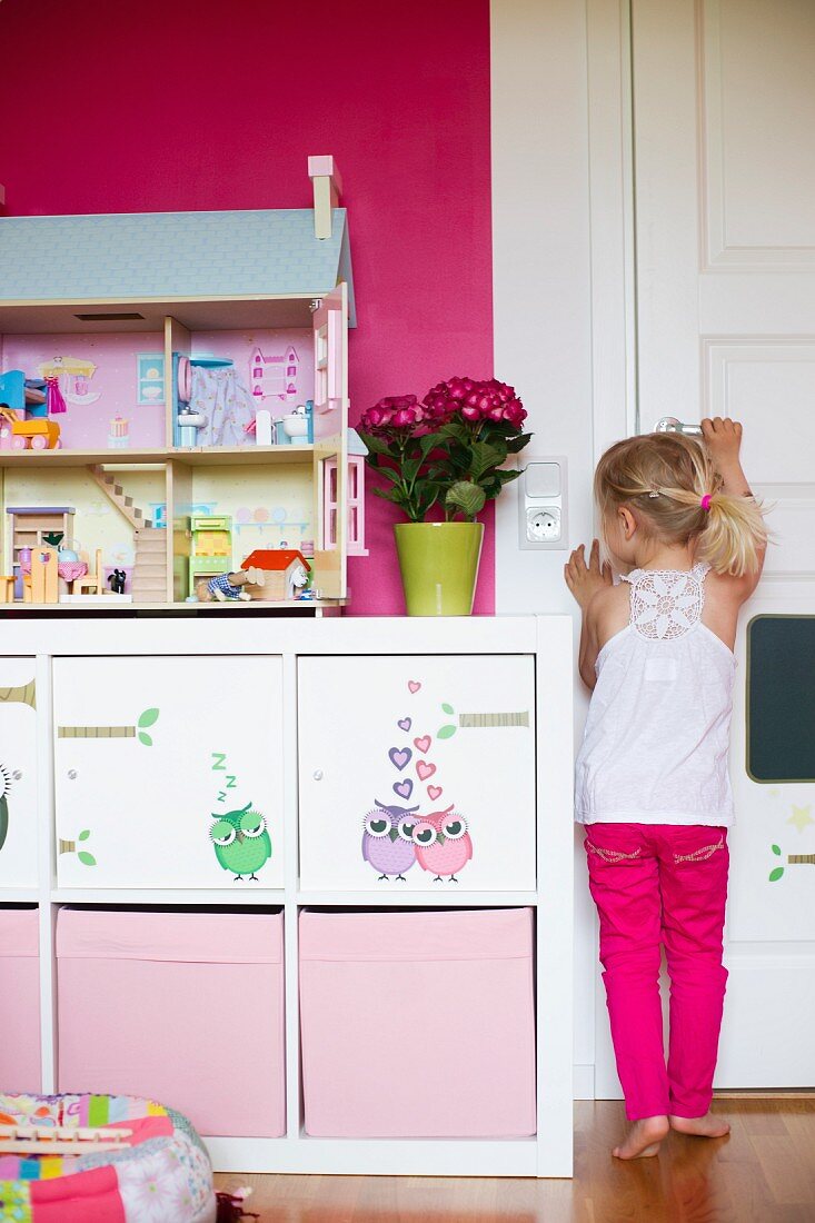 Girl next to white sideboard with storage boxes in compartments and dolls' house on surface against hot pink wall