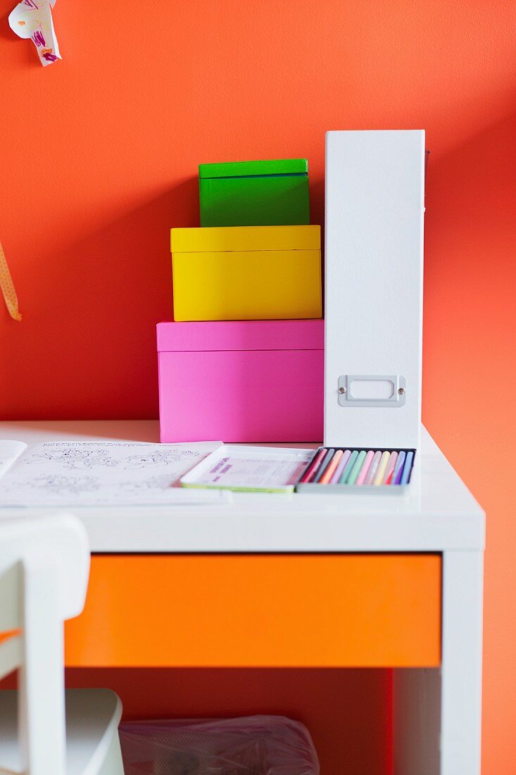 Gestapelte Aufbewahrungsschachteln aus farbigem Karton und weisser Ordner auf Schreibtisch vor orangeroter Wand